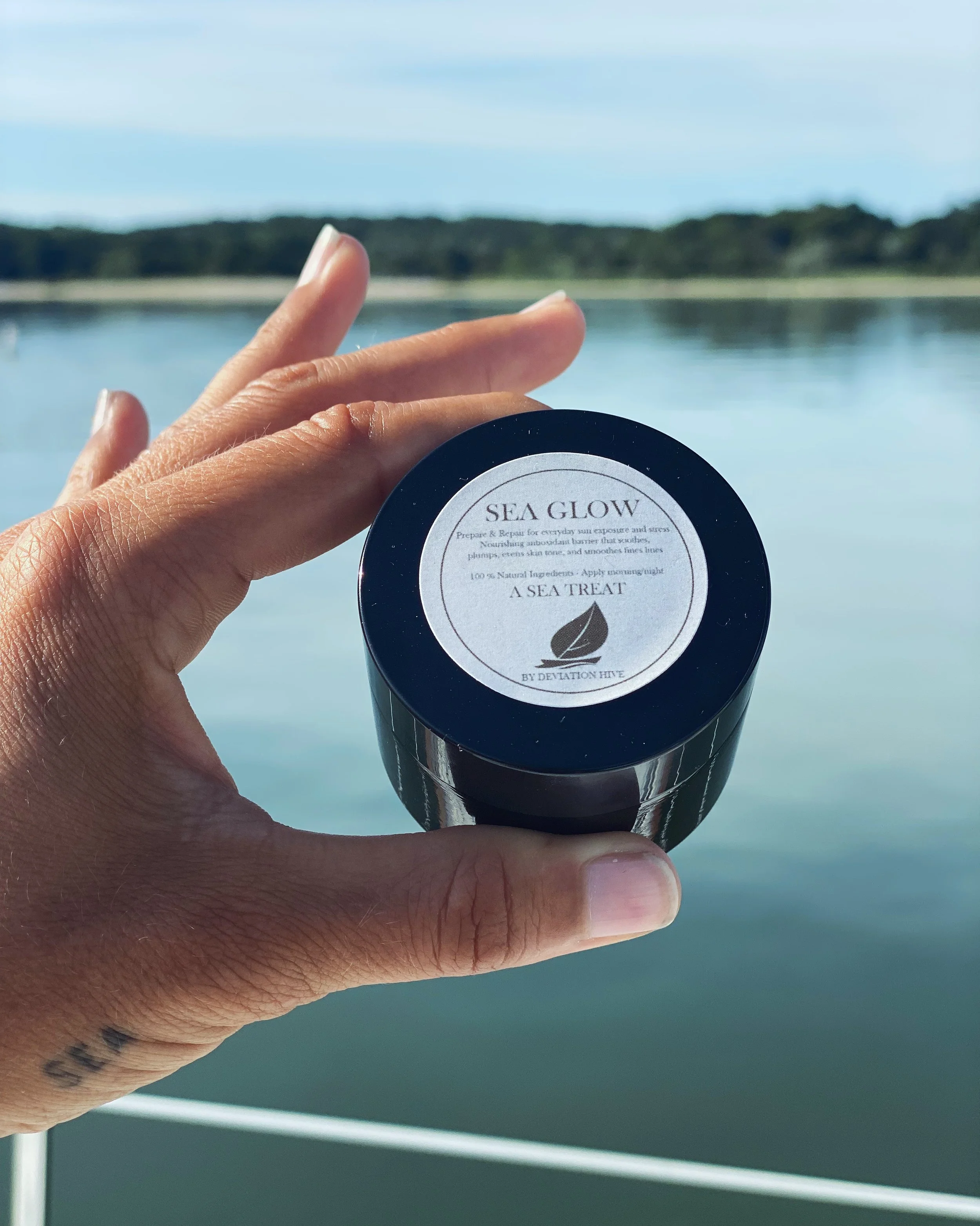 Close-up of a hand holding a container with the mild UV-protecting natural skin care balm 'Sea Glow' near a body of water with trees in the background during daytime.
