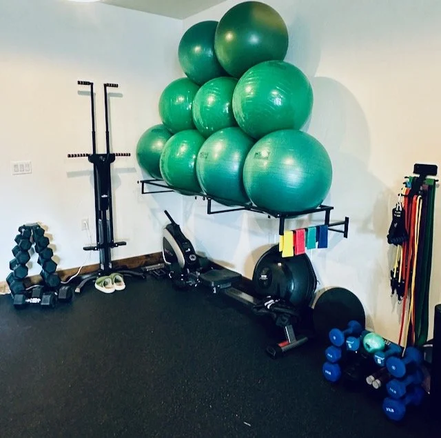 A trainer and a client standing in a gym next to a wall with a big red circle that says 'DO'. The trainer is facing the client with arms at sides giving instruction, and the client is holding a kettlebell in front of him. 