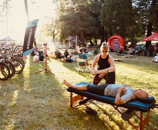 woman stretching a man on a massage table outside, surrounded by bicycles and people sitting in chairs