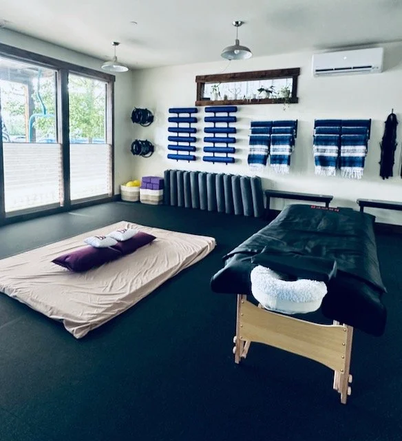 Gym, physical therapy, wellness or yoga studio with exercise equipment and wall-mounted accessories. View of a Thai massage/Stretch Therapy floor mattress with two larger purple pillows, and massage table with head rest and sauna blanket on top.
