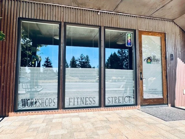Gym, physical therapy, yoga studio with glass windows displaying the words "Workshops," "Fitness," and "Stretch," and a door with a sign that reads "Mellness- Whole Body Wellness." There is an Open sign in the window, and a door mat on a brick porch.