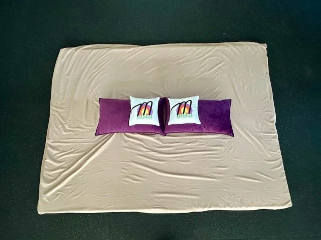 Overhead view of a Thai massage/Stretch Therapy mattress with two larger purple pillows and two smaller white pillows with the Mellness logo printed on them on top of a dark gym floor.