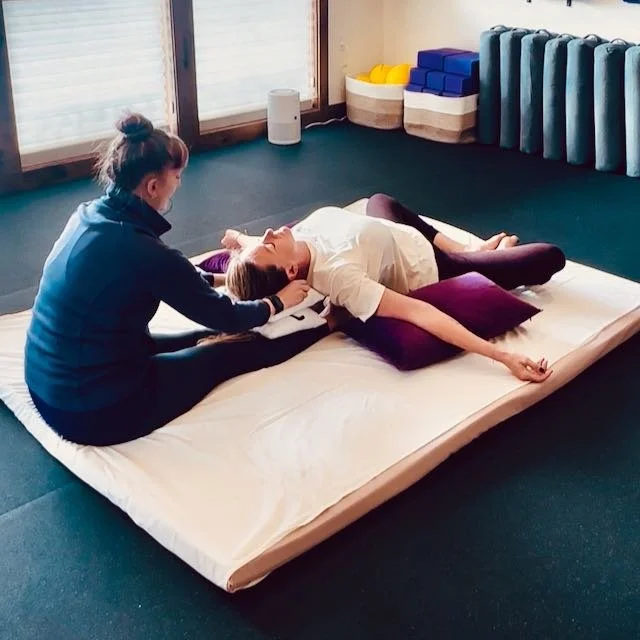 A female therapist performing stretches with one foot on the woman model's sacrum, left had of the therapist on left ankle of the model on on a Thai massage mattress on the floor with pillows and candle around.