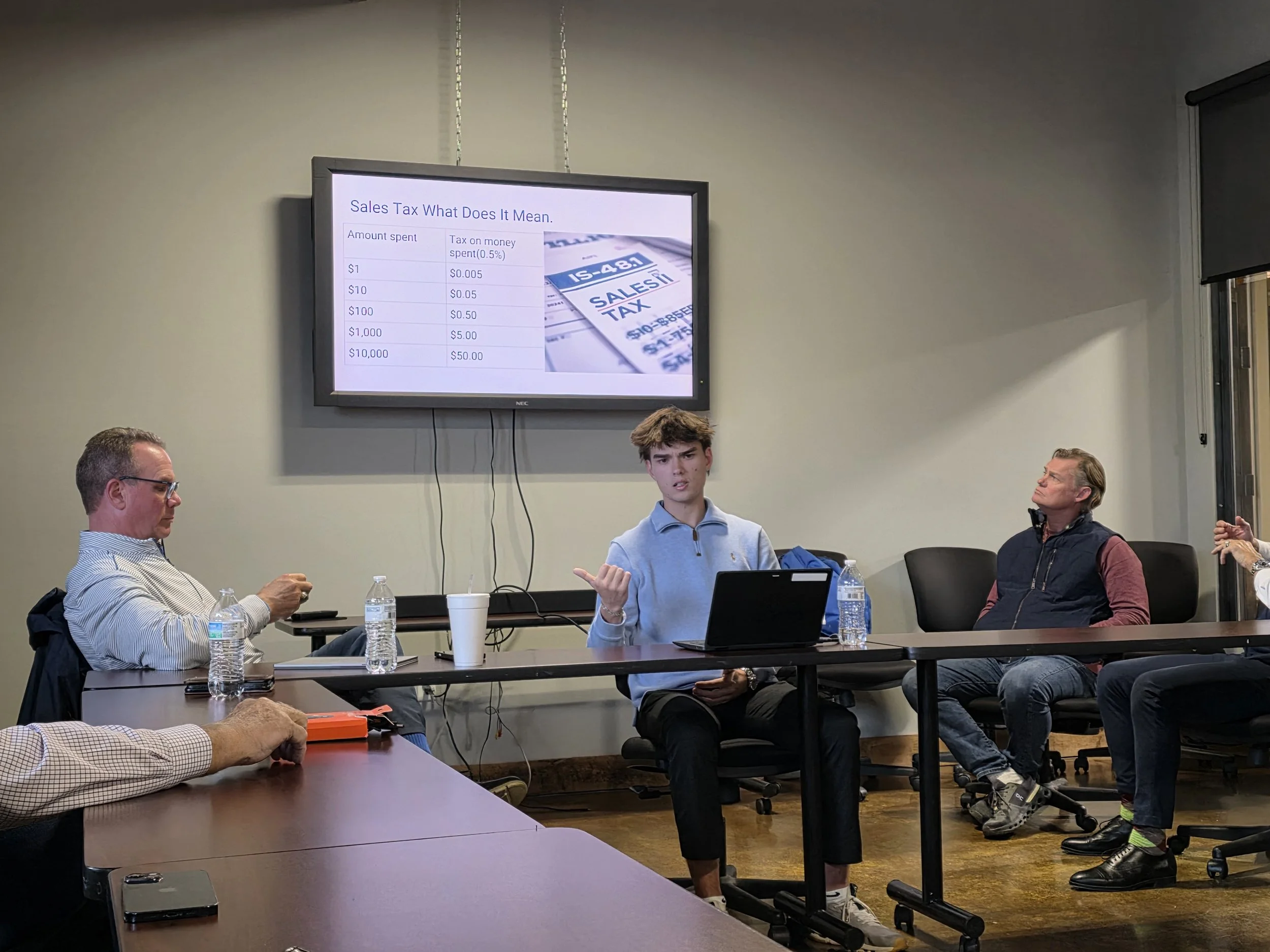 A Northland CAPS student speaks to business professionals during a presentation about sales tax, with a slide showing monetary amounts and corresponding tax displayed on a wall-mounted television.