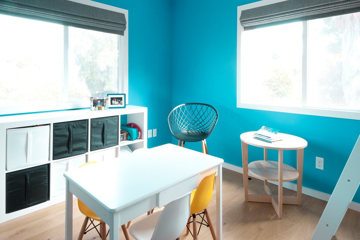 Bright blue children's playroom with window valances, a white table with yellow and white chairs, and a white shelf with black and white storage cubes, a small framed photo, and a picture frame on top. There is a black wire chair and a small wood side table with books next to the windows.