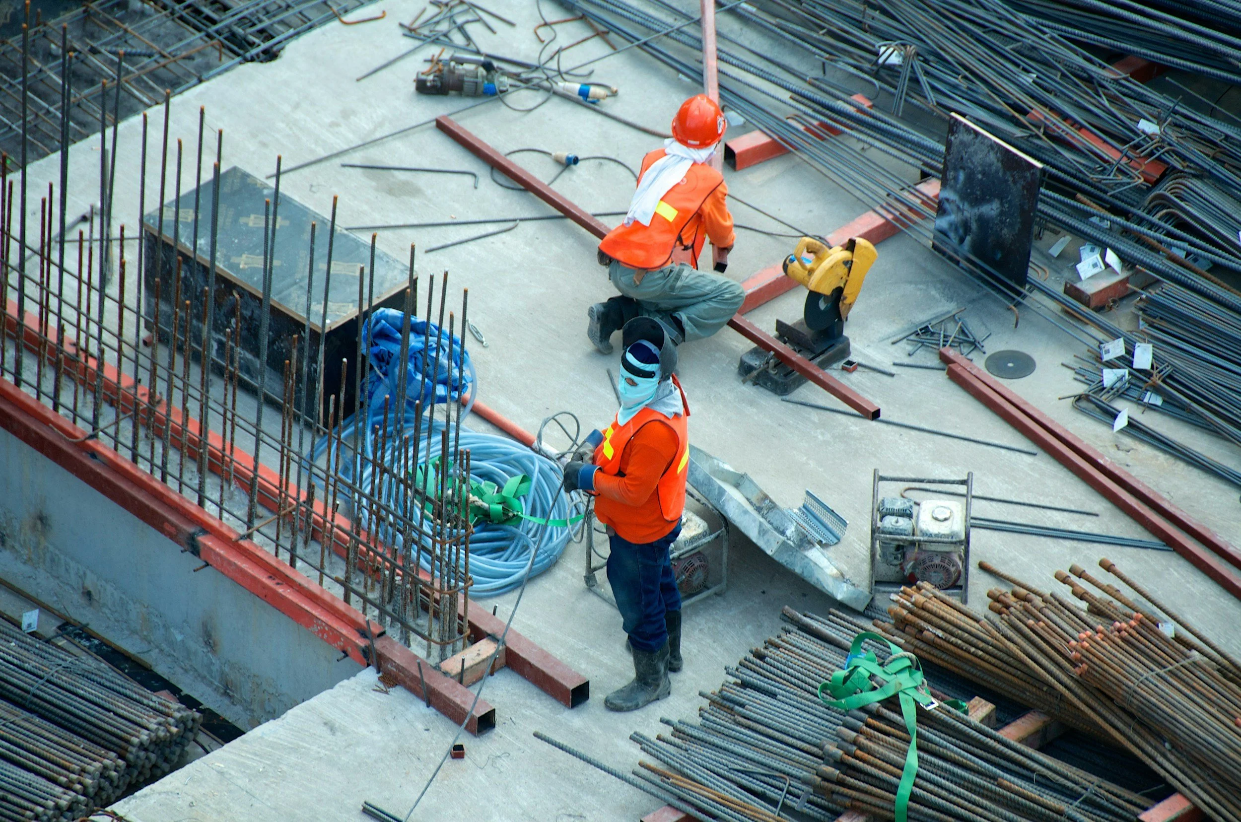 Construction workers wearing orange safety vests, helmets, and masks working on a building site with steel rebar, tools, and construction materials scattered around.