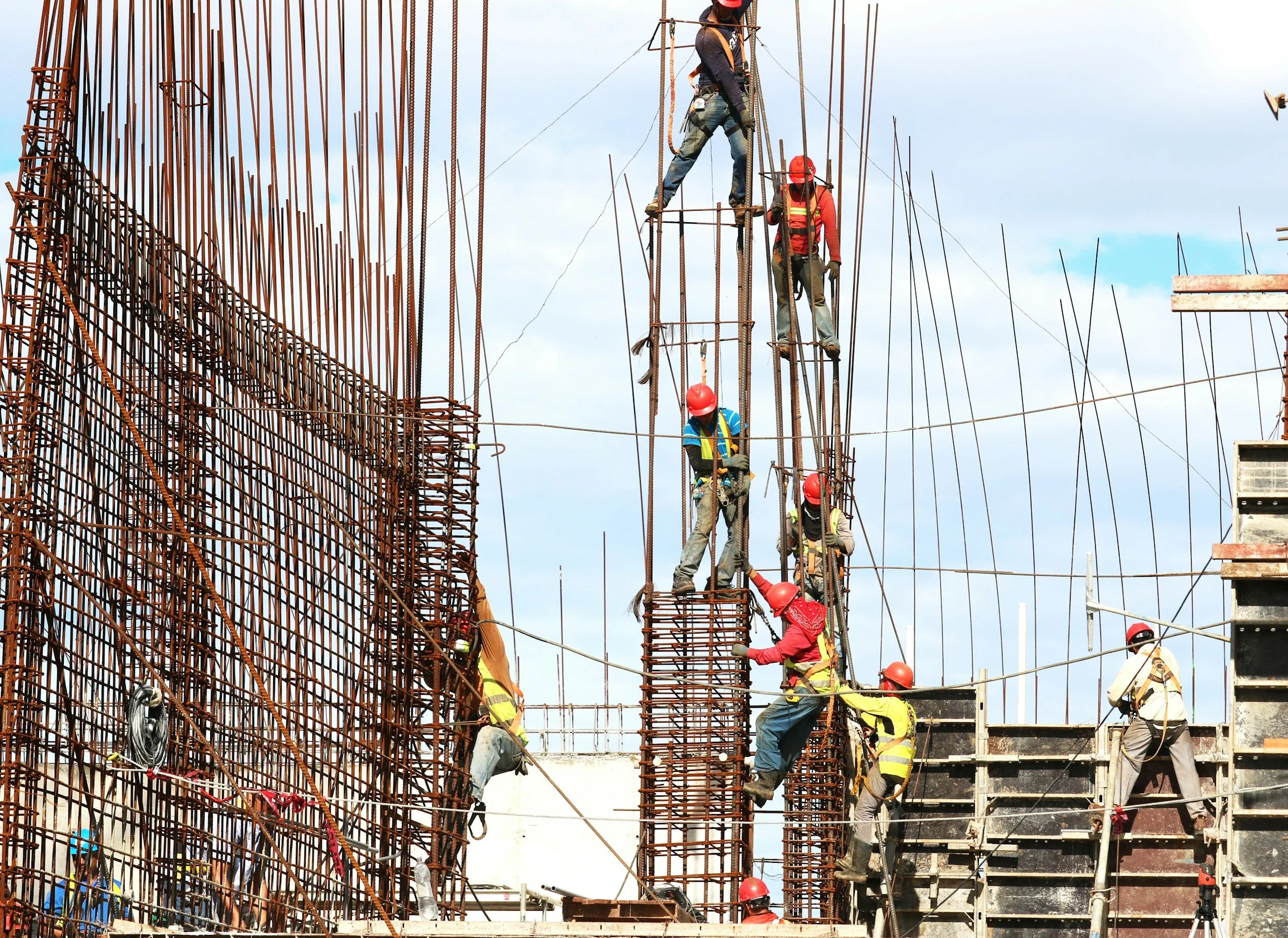 Construction workers wearing safety helmets and harnesses working on a steel reinforcement structure at a building site under a blue sky.