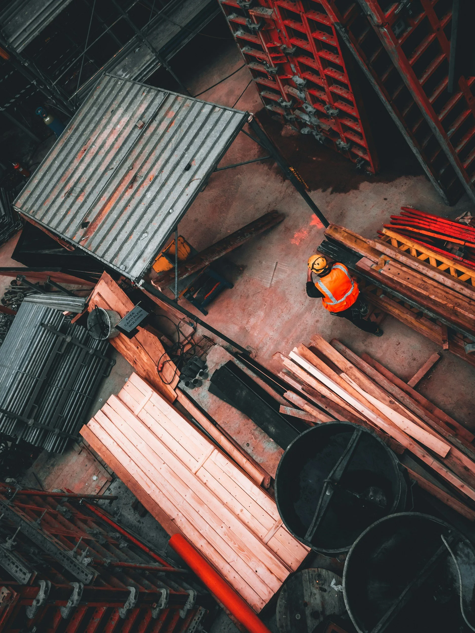 A worker in a yellow helmet and orange safety vest standing in a construction site, surrounded by stacked wooden planks, metal pipes, and construction materials.