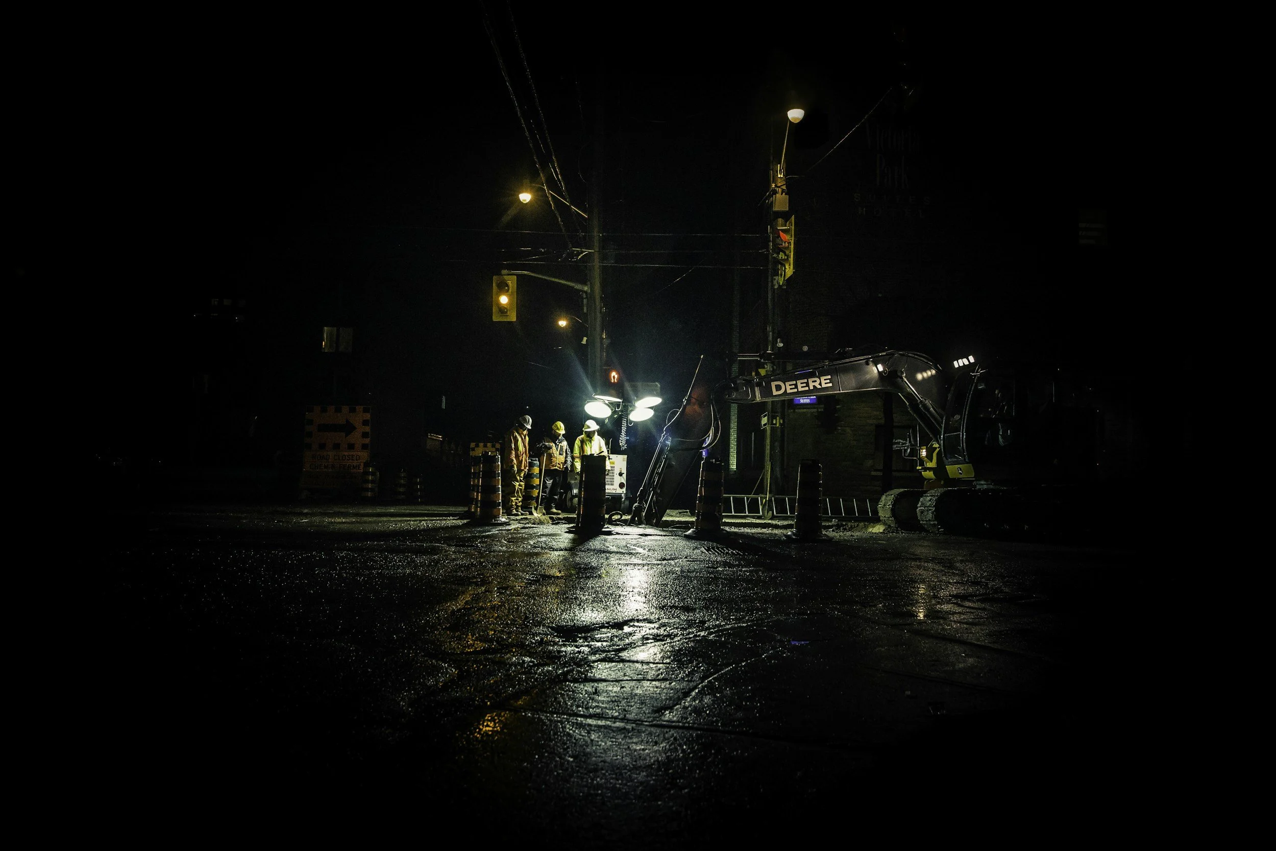 Nighttime construction scene with workers, a Deere excavator, traffic lights, and wet ground reflecting lights.