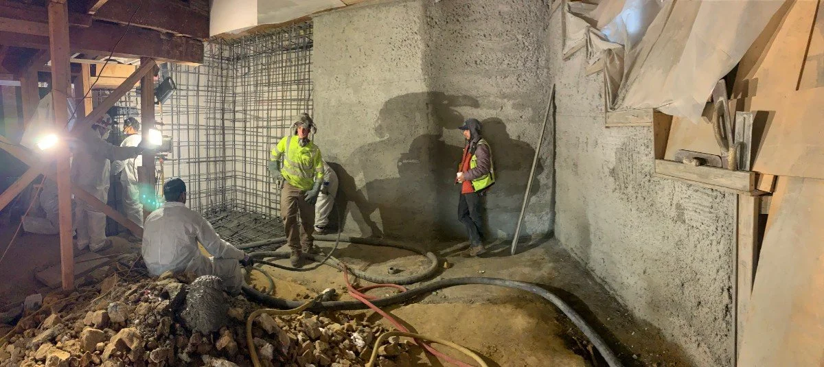 Construction workers in safety gear working inside a building with concrete walls, rebar, and scattered construction materials.