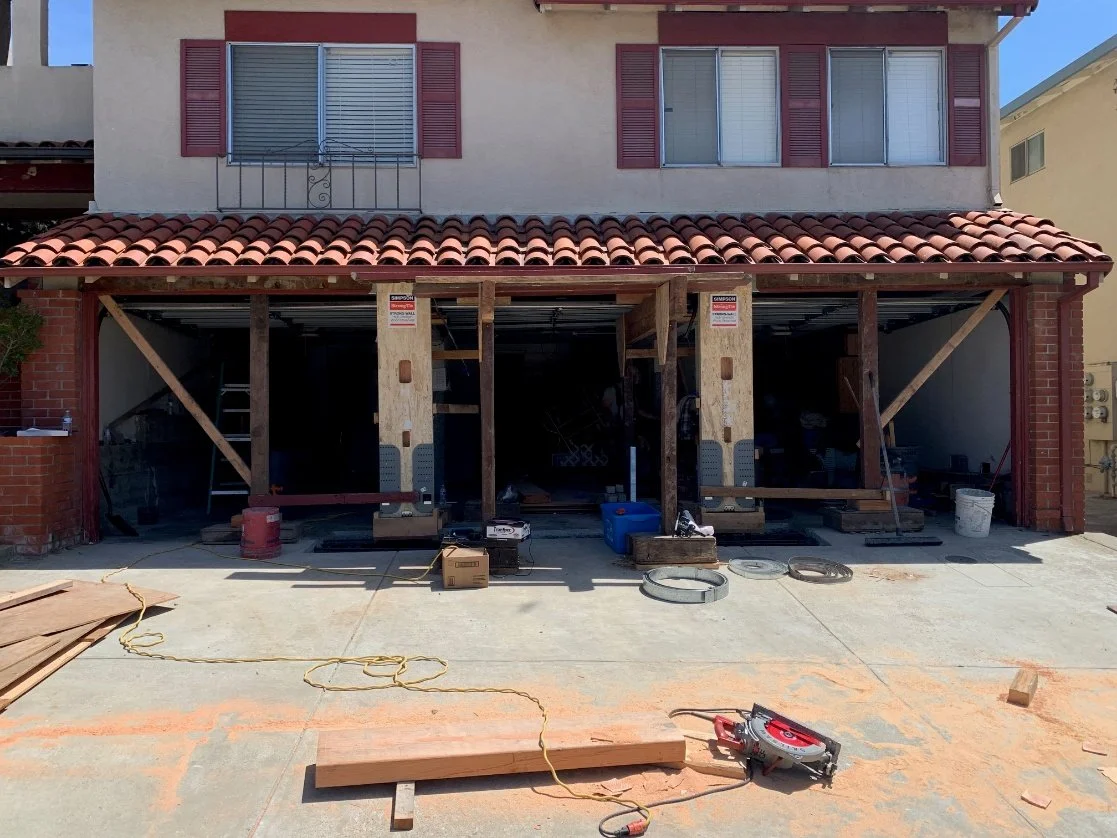 House undergoing renovation with construction materials and tools on the driveway, exposed wooden framework in the garage, and upper floor windows with closed shutters.