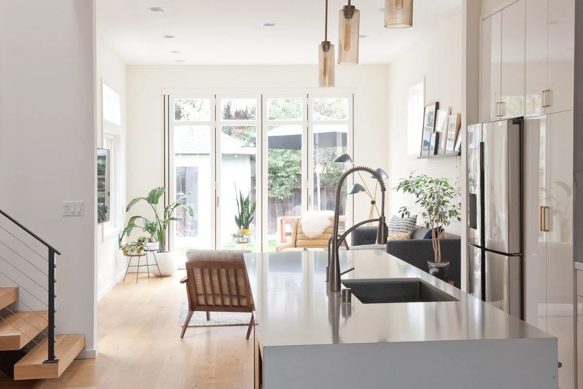 Bright, modern kitchen with a white island, stainless steel sink, hanging pendant lights, and large windows with a view of a backyard with trees and outdoor furniture. Indoor plants and a cozy seating area are visible.