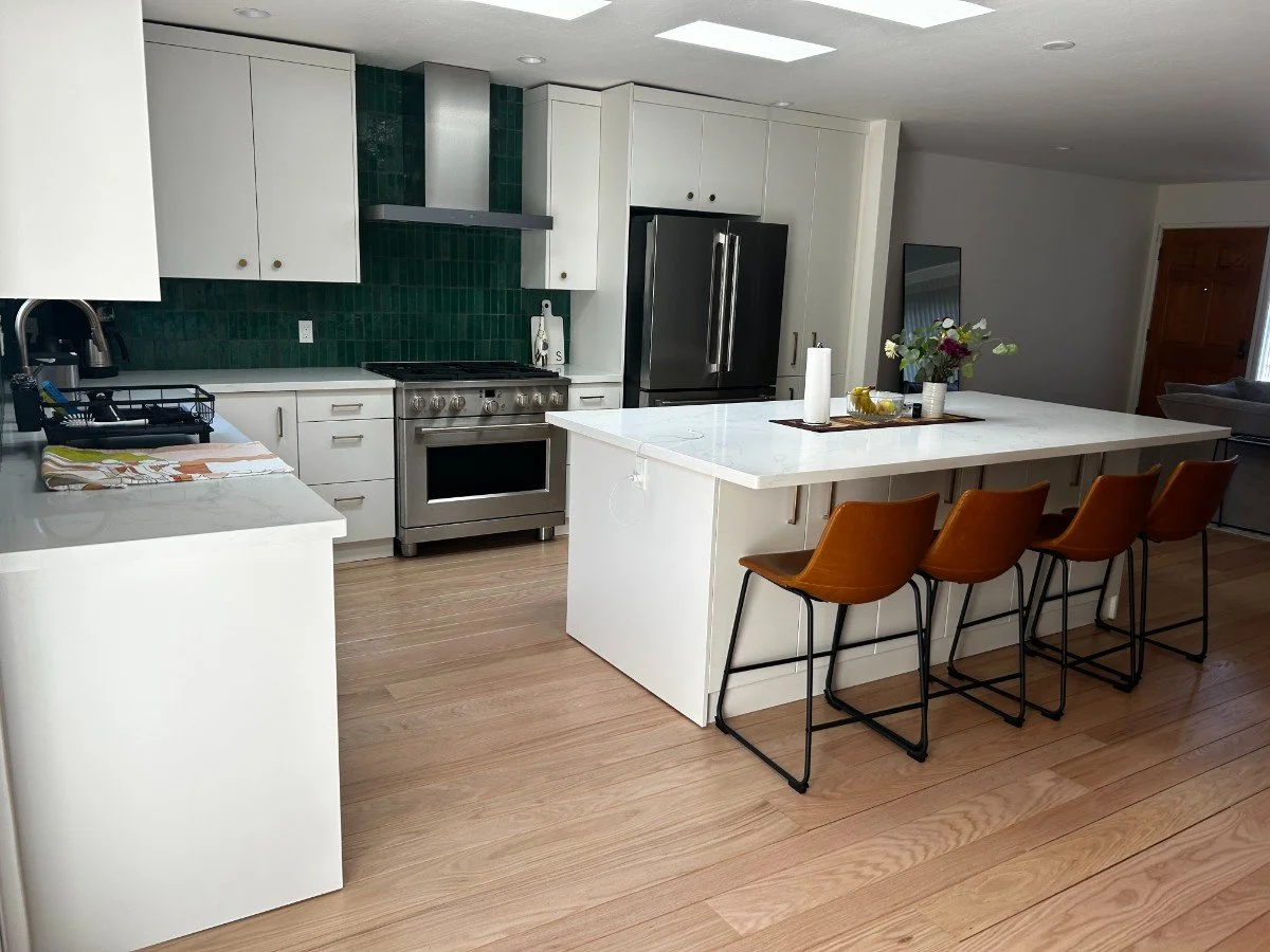 Modern kitchen with white cabinets, a gray stove, and a large stainless steel refrigerator. There is a white kitchen island with a vase of flowers, paper towels, and fruit. Four brown bar stools are lined up along the island. The floor is light wood,