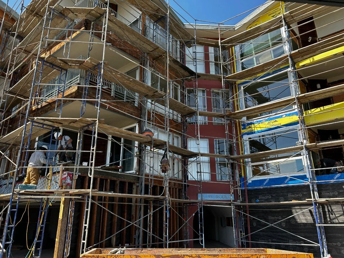 Construction workers building or renovating a multi-story building, with scaffolding covering the front of the structure and construction materials visible.