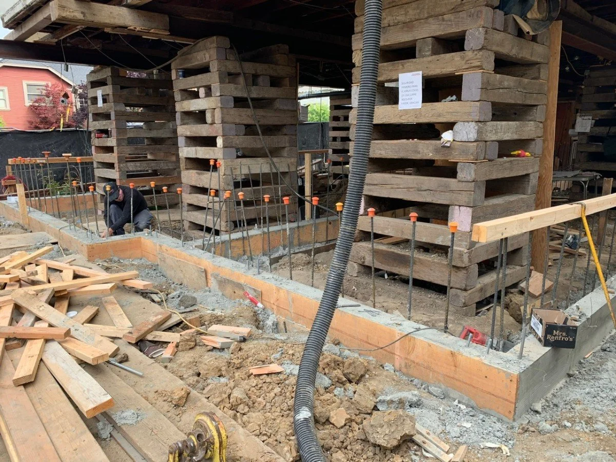 Construction site with a worker kneeling on the ground, working on the foundation, surrounded by wooden and metal framing and stacked wooden pallets.