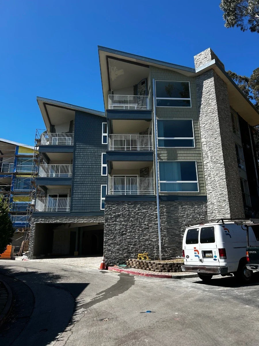 Modern multi-story apartment building with balconies, stone and siding facade, and a driveway with construction cones and a parked white van.