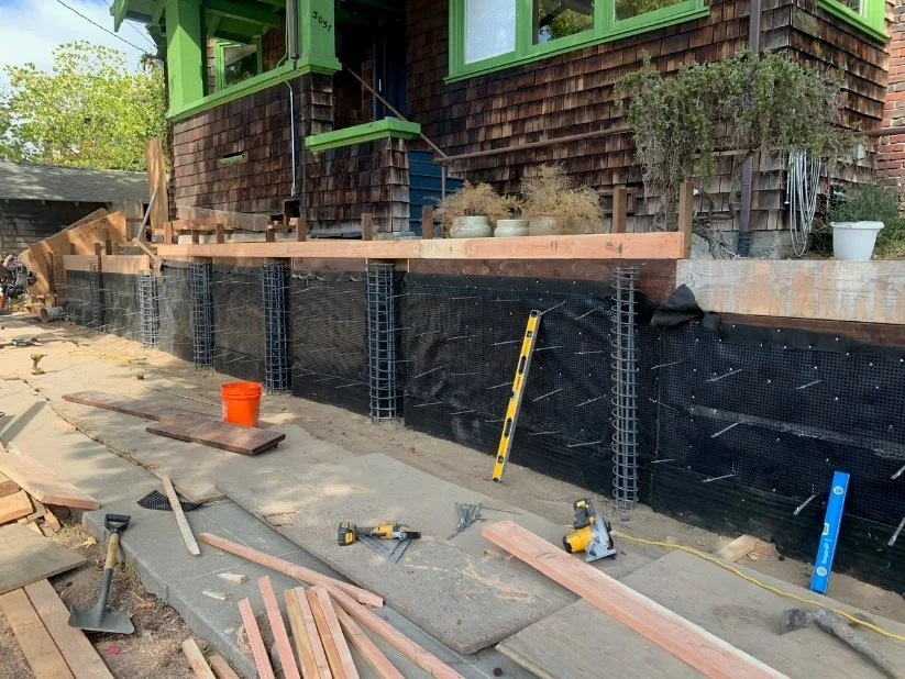 Construction site with foundation work, showing rebar, black waterproofing membrane, and wooden formwork for a building. Tools and materials are scattered around.