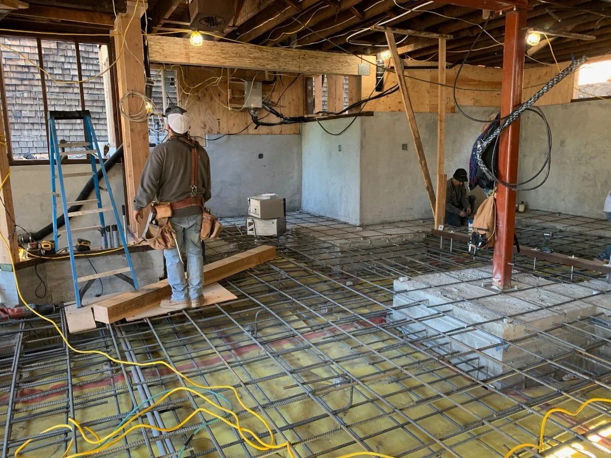 Interior of a building under construction, showing exposed wooden framing, concrete walls, electrical wiring, and people working on the floor with metal rebar and tools.