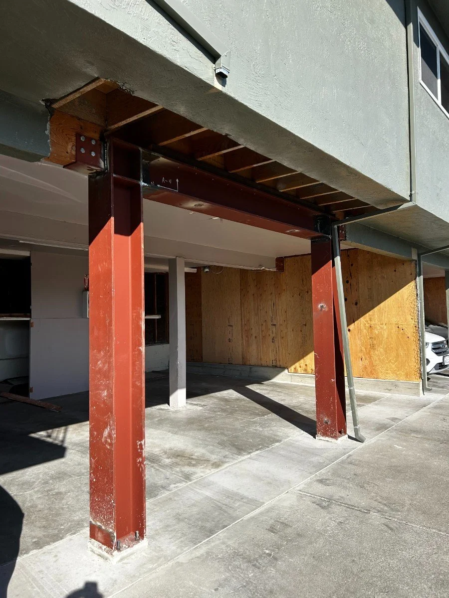 Construction site of a building showing reinforced steel beams and support columns, with wooden framing and concrete pavement.