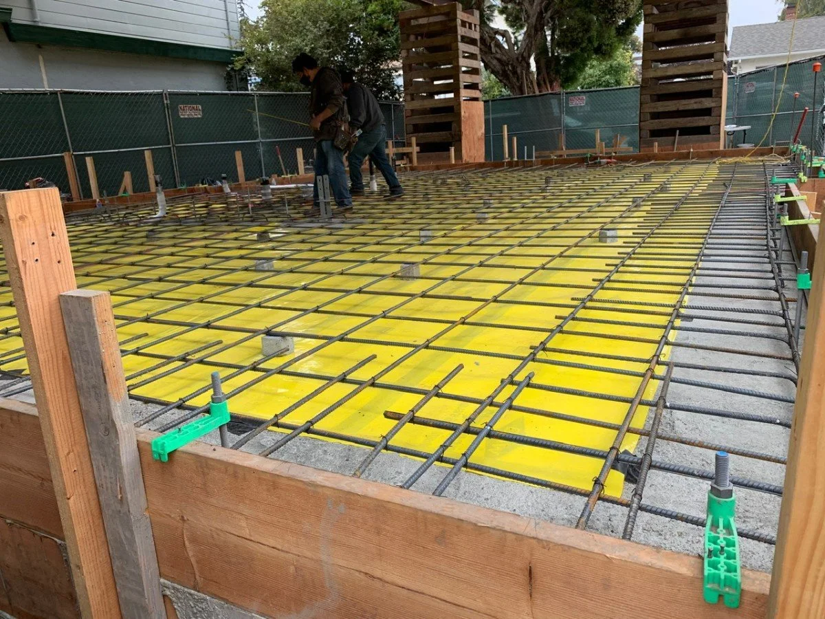 Construction workers laying rebar and yellow plastic sheets for a concrete slab at a residential building site. The area is surrounded by green fencing and wooden formwork.