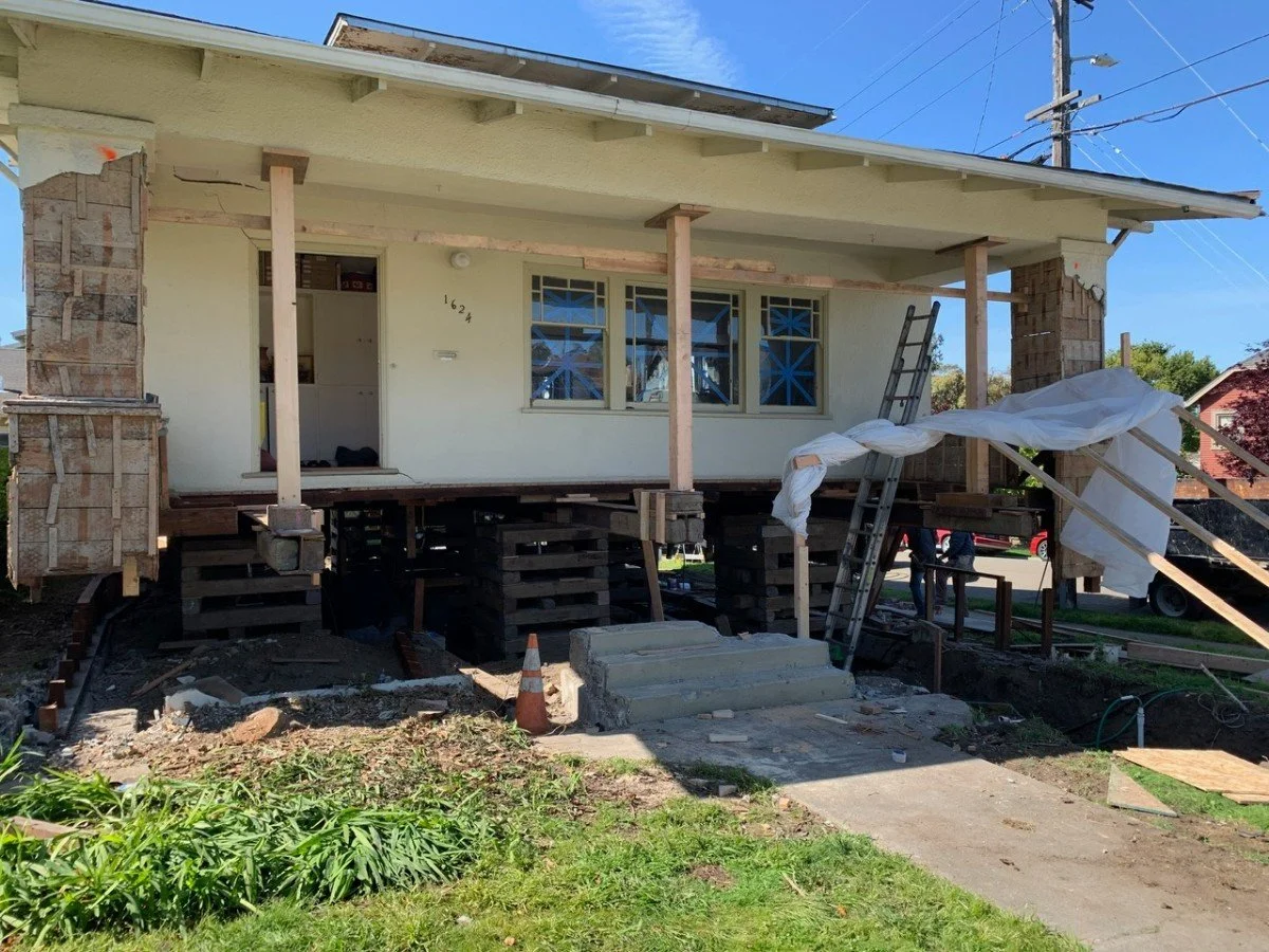 A house under renovation with its foundation elevated and supported by concrete blocks. The exterior walls are partially finished, with some areas showing brickwork and others covered with light-colored siding. Scaffolding and ladders are present aro
