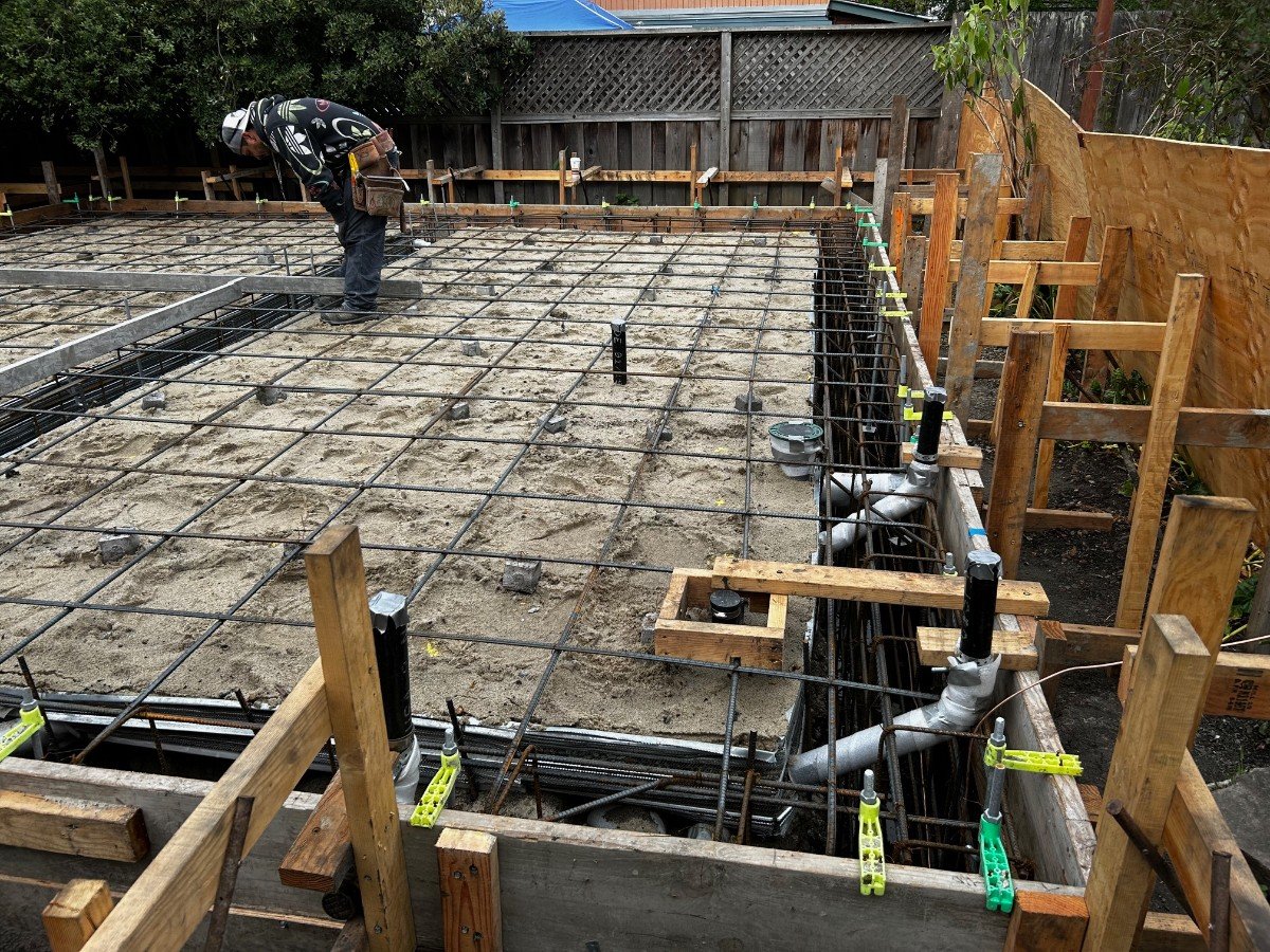 Construction site with rebar grid and plumbing pipes for building foundation, worker inspecting it during daytime.