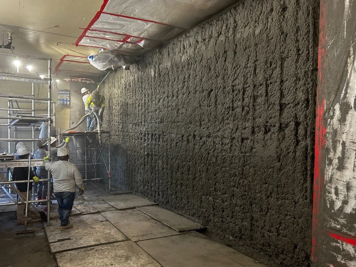 Construction workers applying spray concrete on a large vertical wall inside a building, with scaffolding and construction materials present.