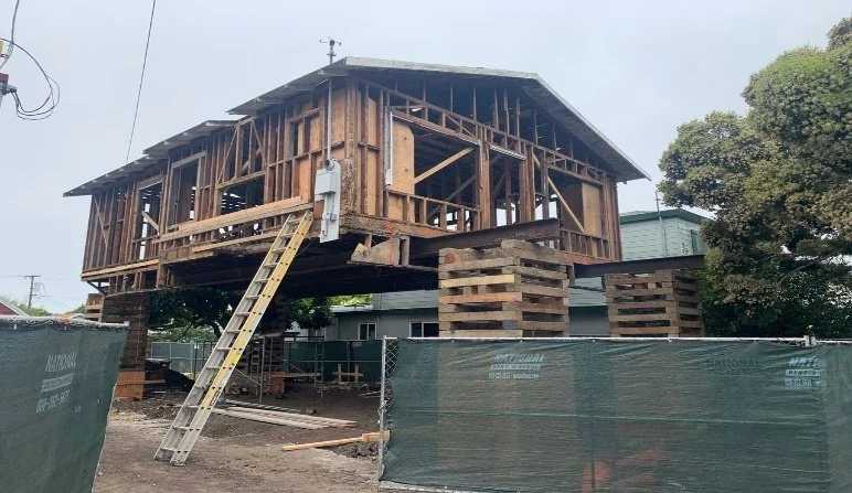 Wooden house under construction lifted on stilts, with a ladder leaning against it, surrounded by construction fencing and nearby trees.