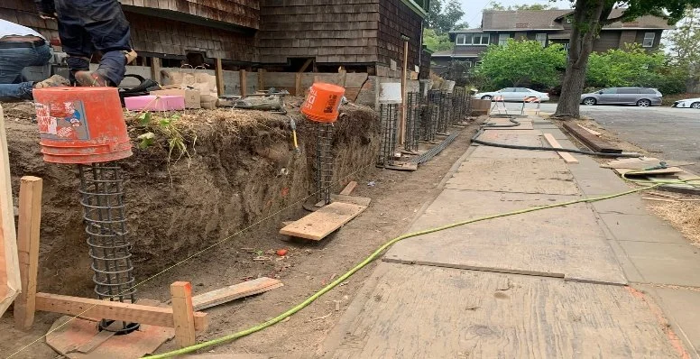 Construction site with rebar columns, wooden formwork, and a partially torn sidewalk. A worker is seen near a pile of construction materials, and orange buckets are placed on the rebar.