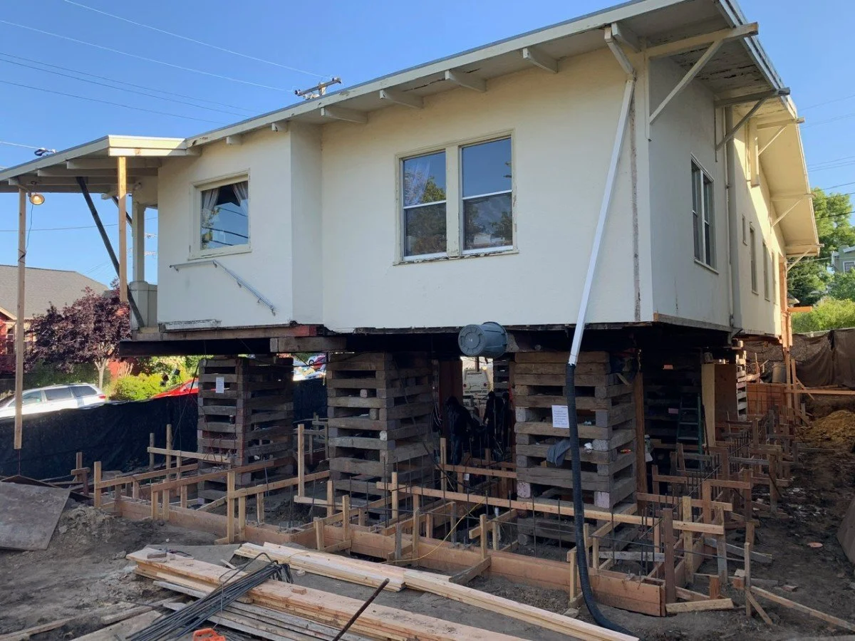 A house elevated on wooden pallets and supports during construction or renovation, with construction materials and framing around the site, under a clear blue sky.