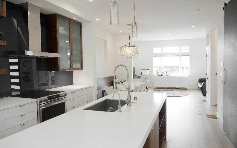 Modern open-concept kitchen with white island, stainless steel appliances, and contemporary lighting, adjacent to living area with a large window, sofa, and decorative rug.