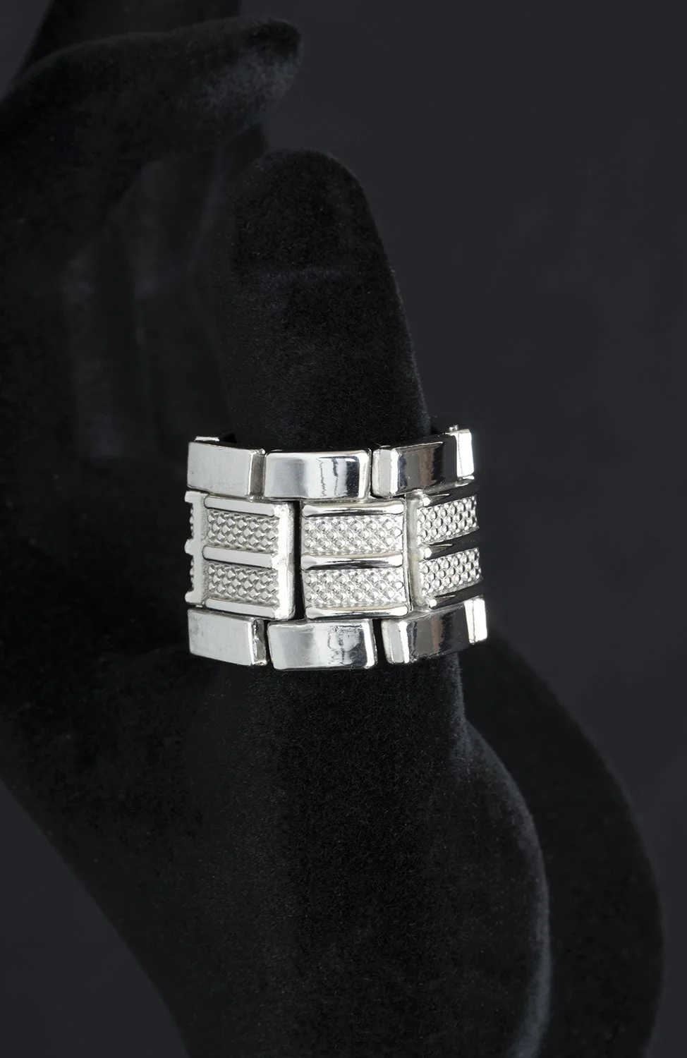Close-up of a silver ring with a geometric pattern, worn on a finger covered with black fabric, against a dark background.