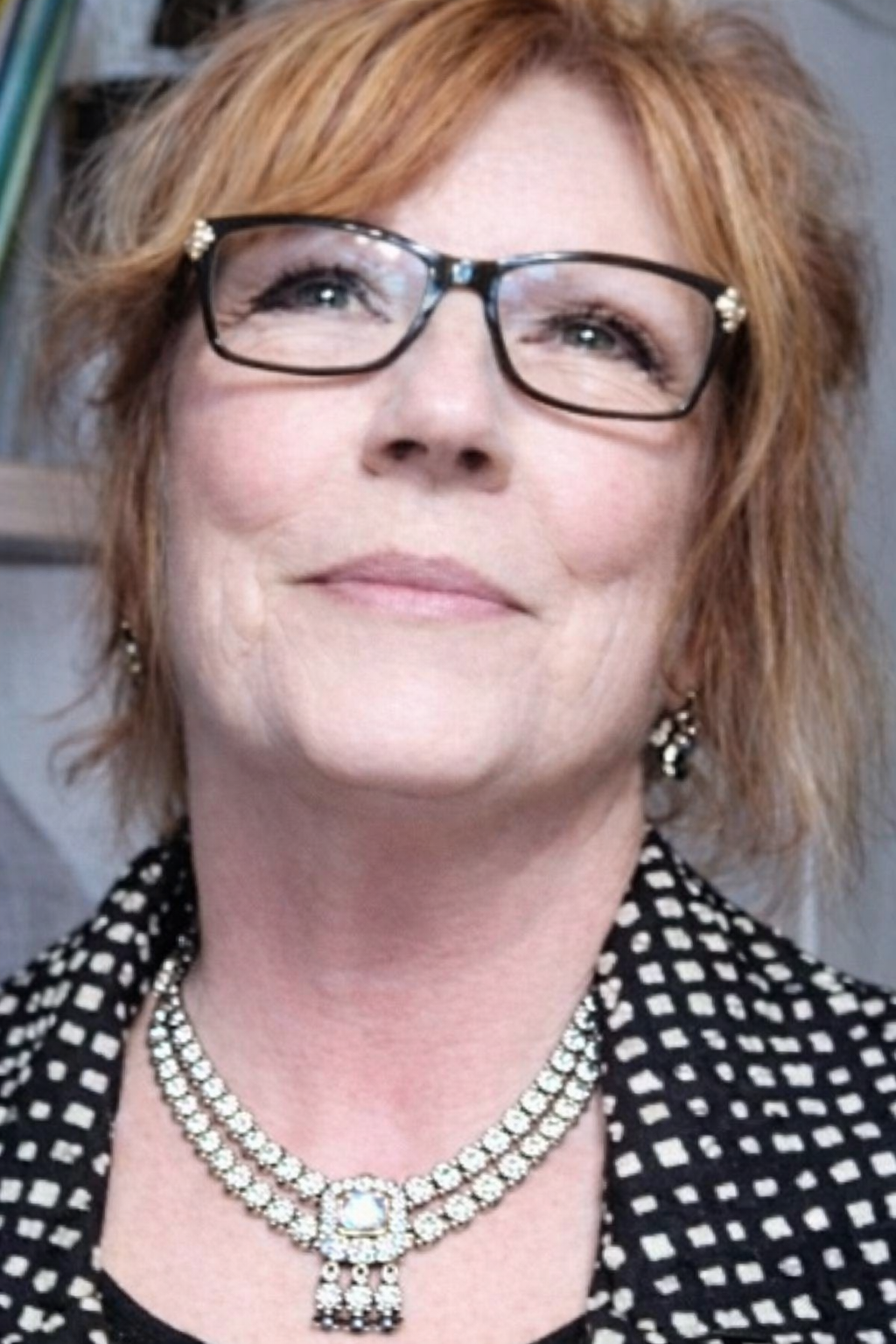 A woman with short, wavy, red hair and glasses, wearing a black-and-white checkered top, a layered diamond necklace, and matching earrings, smiling slightly.