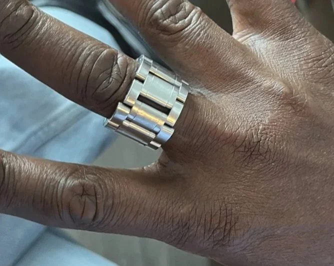 A hand with dark skin wearing a silver ring on the middle finger, and holding a smartphone.