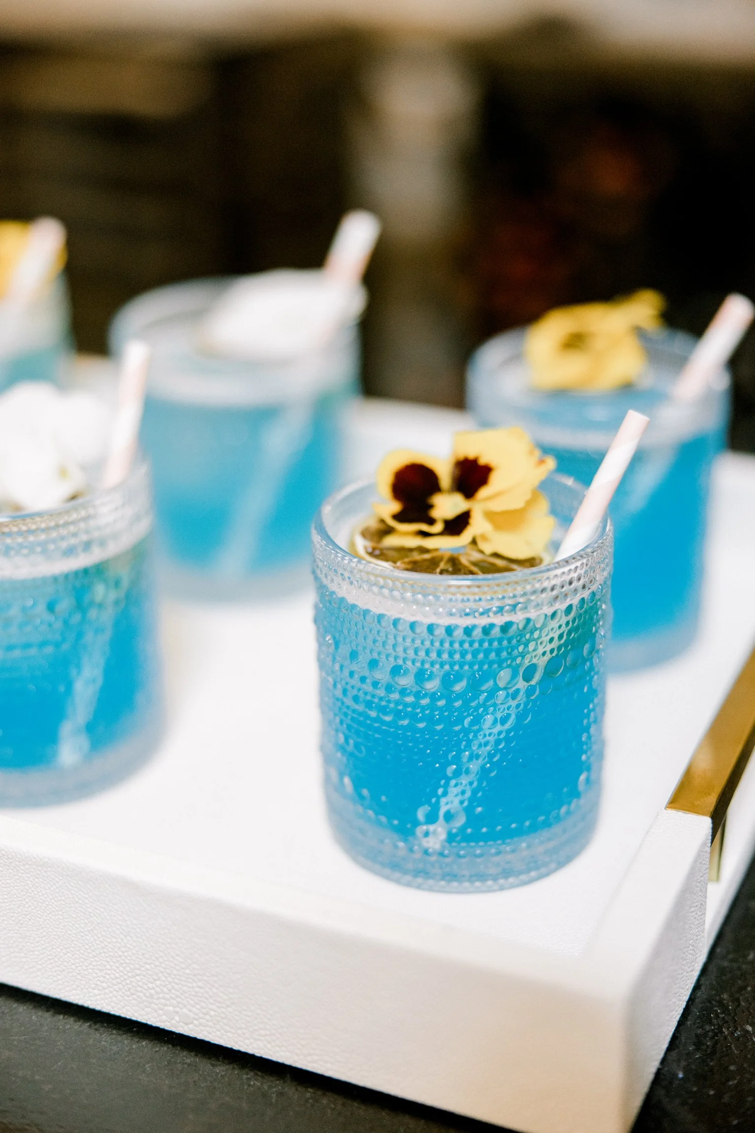 Four blue drinks in textured glasses with white straws and yellow flower garnishes on a white tray.