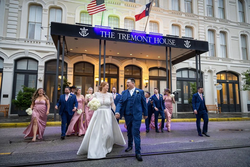 A wedding party walking out of a building called The Tremont House. The bride and groom are at the front, holding hands and smiling, surrounded by bridesmaids in pink dresses and groomsmen in blue suits. The building has American and Texas flags flying above the entrance.