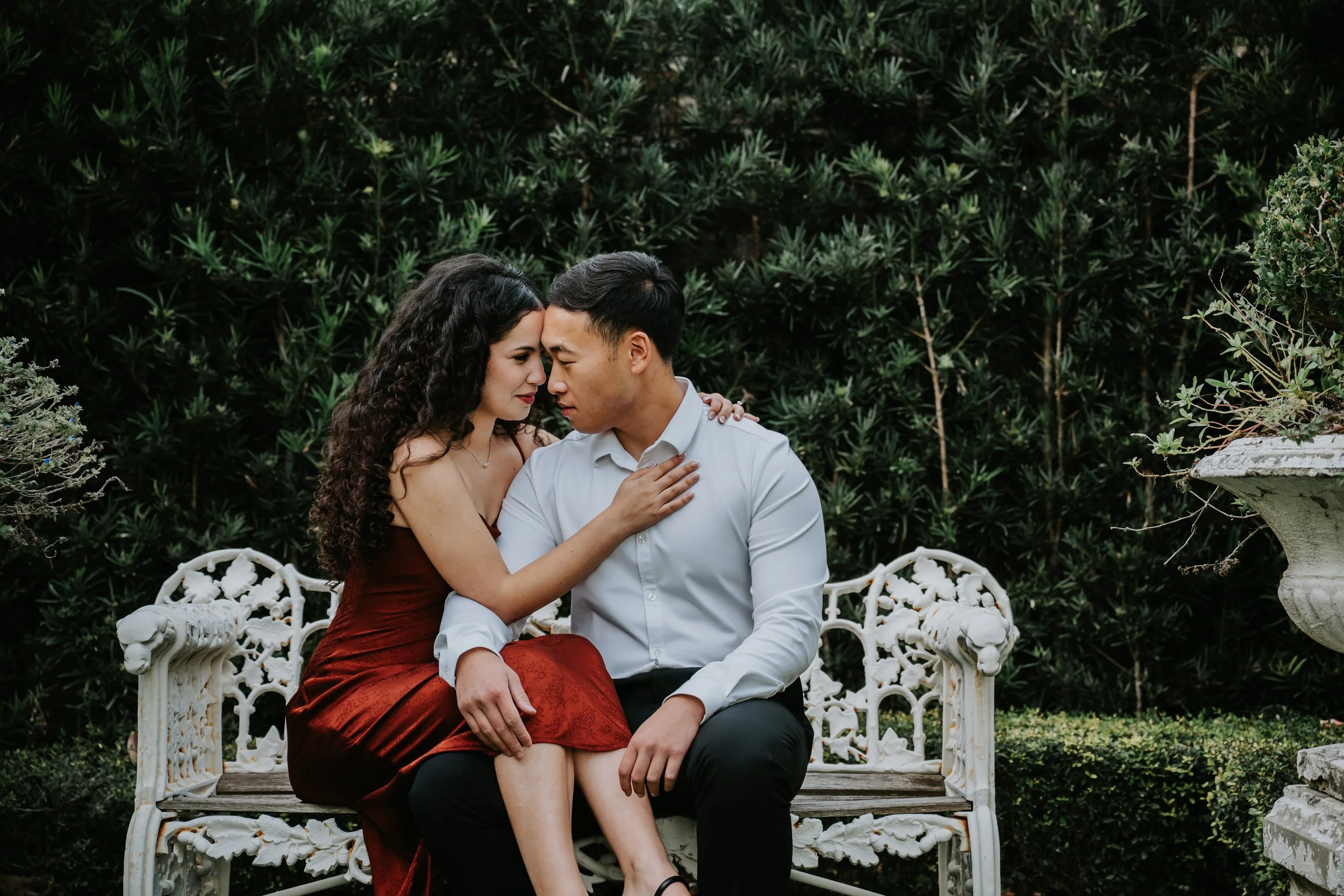 A couple sitting closely on a white ornate garden bench with lush green foliage in the background, with foreheads touching and eyes closed.