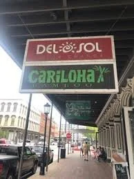 Street view with store signs for Del Sol and Cariloha, sidewalk with pedestrians, parked cars, and buildings