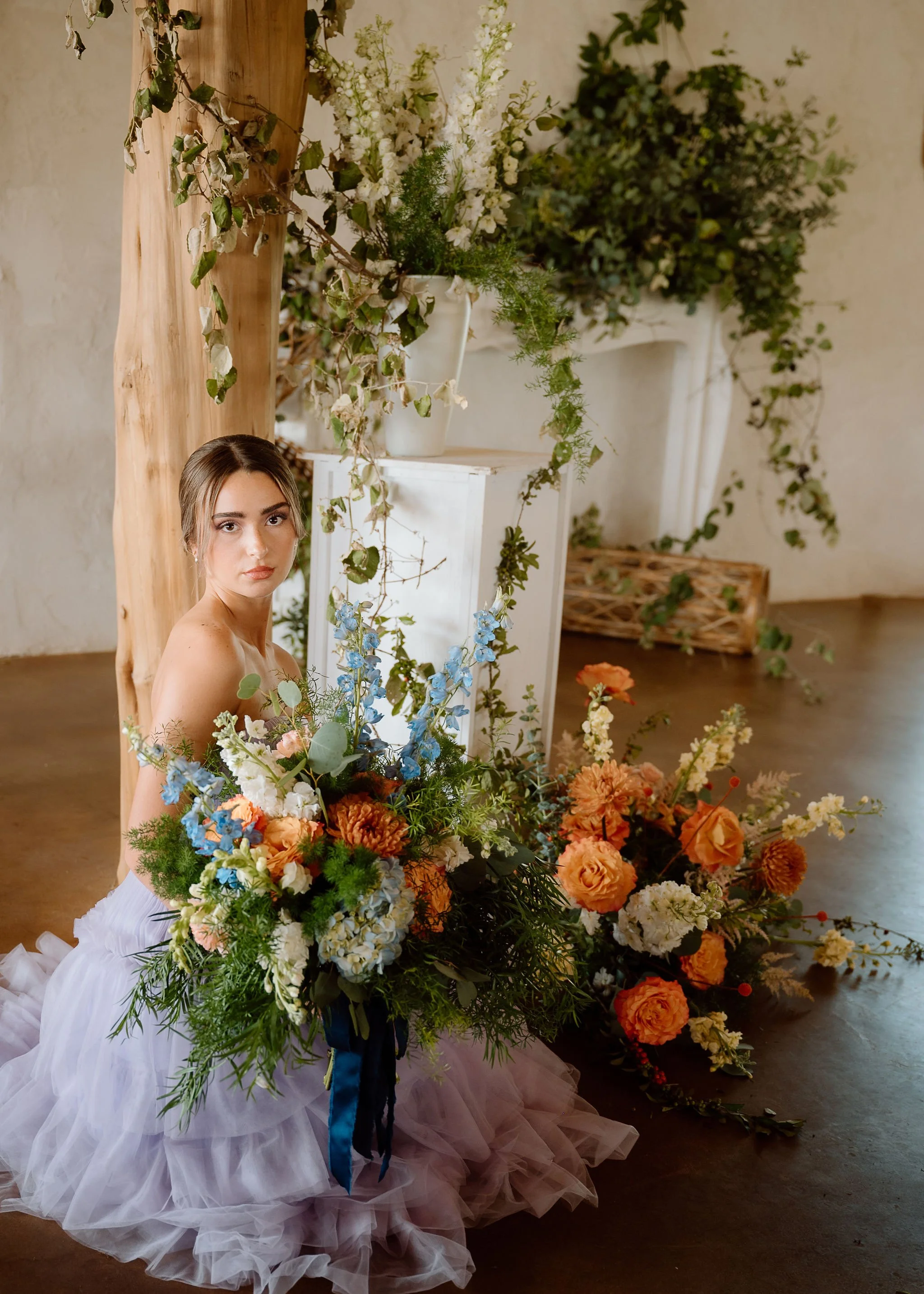 A woman in a white dress surrounded by an abundance of colorful flowers and greenery, with a large floral arrangement in the background and a rustic wooden post.