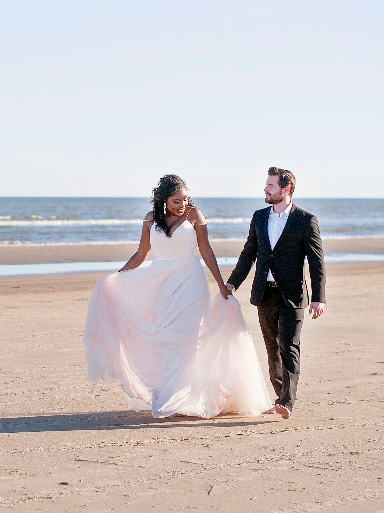 A couple in wedding attire sitting on a sofa on the beach, holding drinks, with a mobile bar trailer nearby, decorated with flowers and tableware.