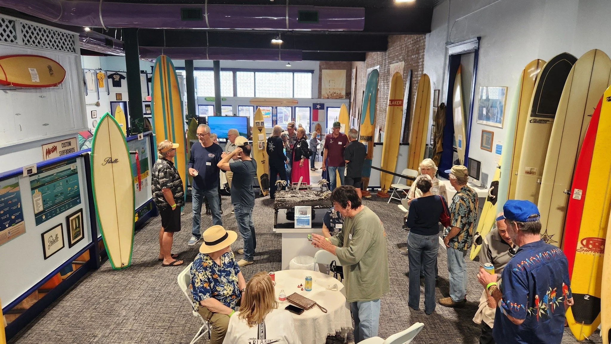 People gathered in a surfing shop or museum, with surfboards displayed on walls and a large flat-screen TV at the back. The scene shows various individuals socializing, some seated at tables and others standing, surrounded by surf-related memorabilia and art.