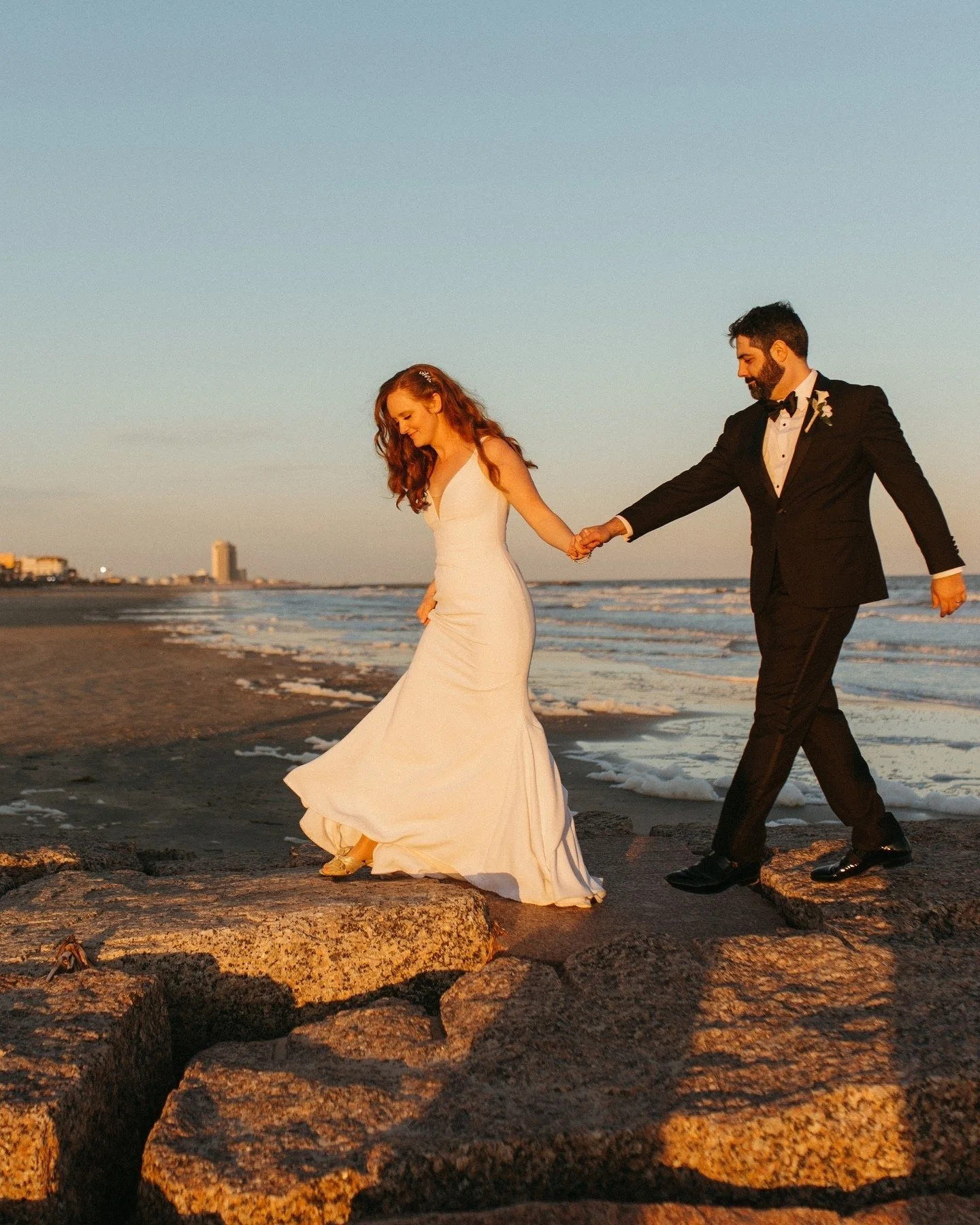 A bride and groom walk hand in hand on a beach at sunset, with the ocean and city buildings in the background.
