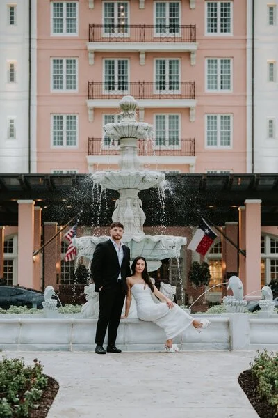 A couple in wedding attire standing in front of a decorative fountain outside a pink apartment building with balconies. The man is in a black suit and the woman is in a white dress.