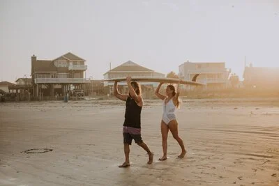 A man and woman walking on the beach carrying a long wooden plank at sunset.