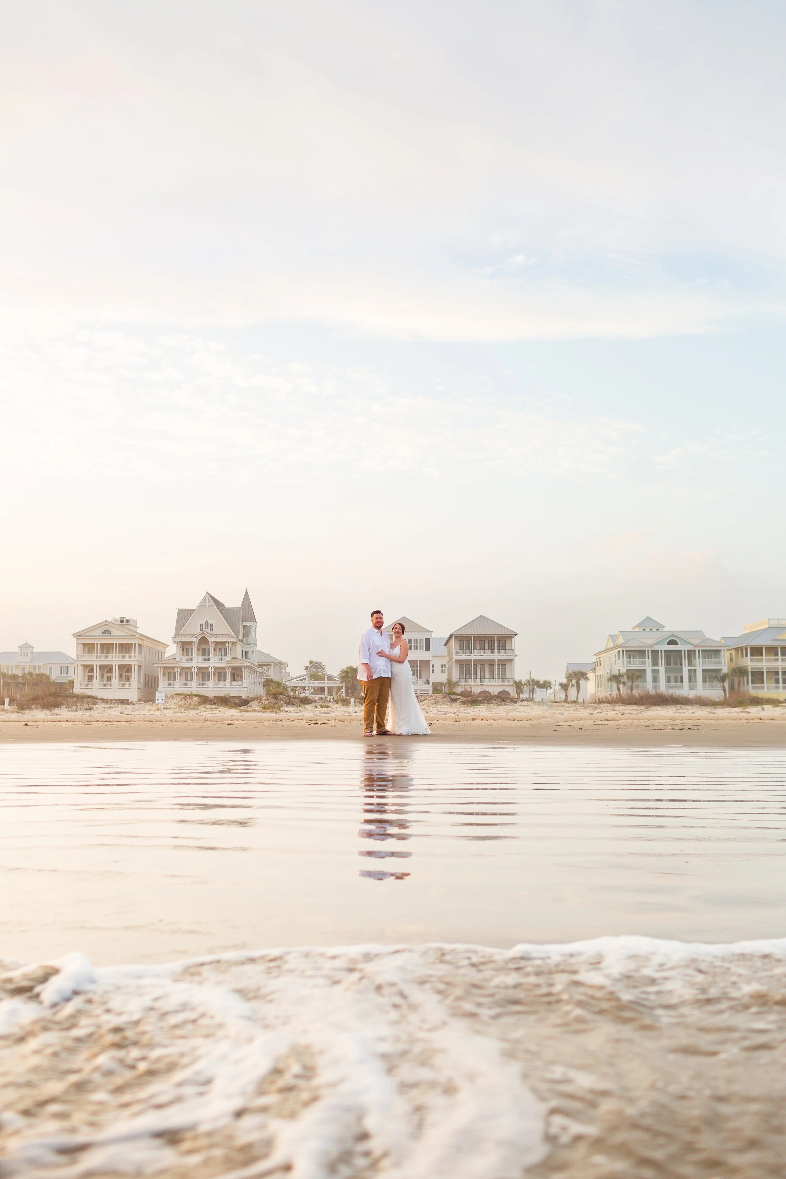 A couple standing on the beach near the water, with large beach houses in the background during sunset or sunrise.