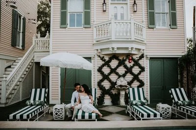 Two people sitting on lounge chairs in front of a pink house with green shutters, decorated with a large black wreath and a white umbrella for shade.