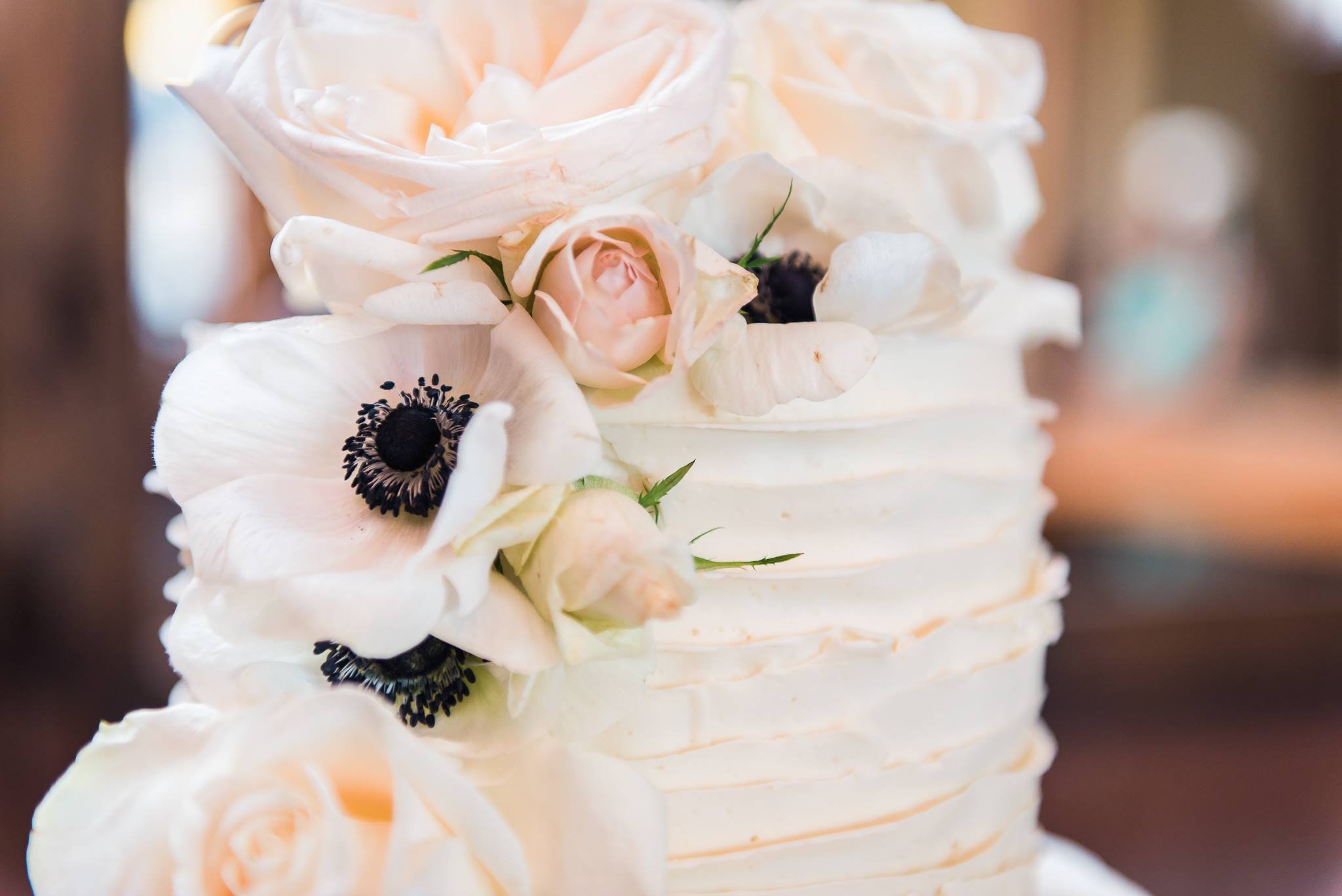 A close-up of a white layered cake decorated with light pink roses, white anemone flowers, and black centers, with smooth frosting and minimal piping details.