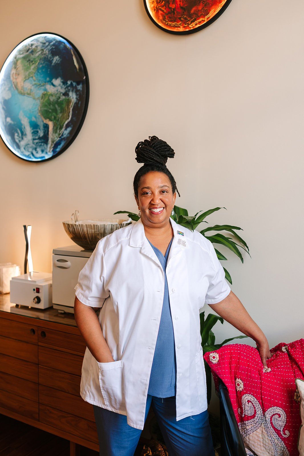 A woman with dreadlocks tied in a bun, smiling and wearing a white medical coat over blue scrubs, standing in a room with artwork and plants.