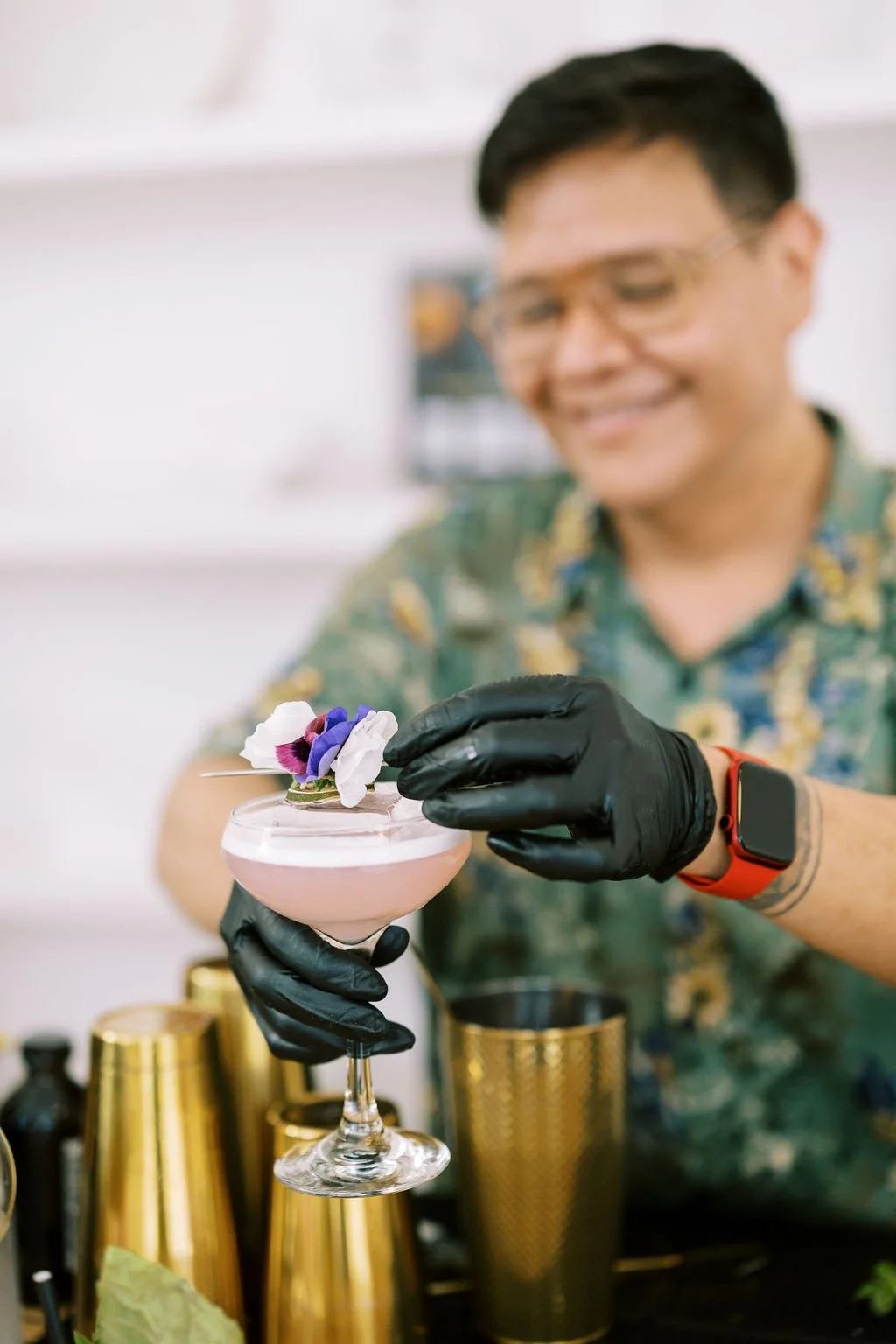 A bartender wearing black gloves and glasses, smiling while garnishing a pink cocktail with edible flowers at a bar.
