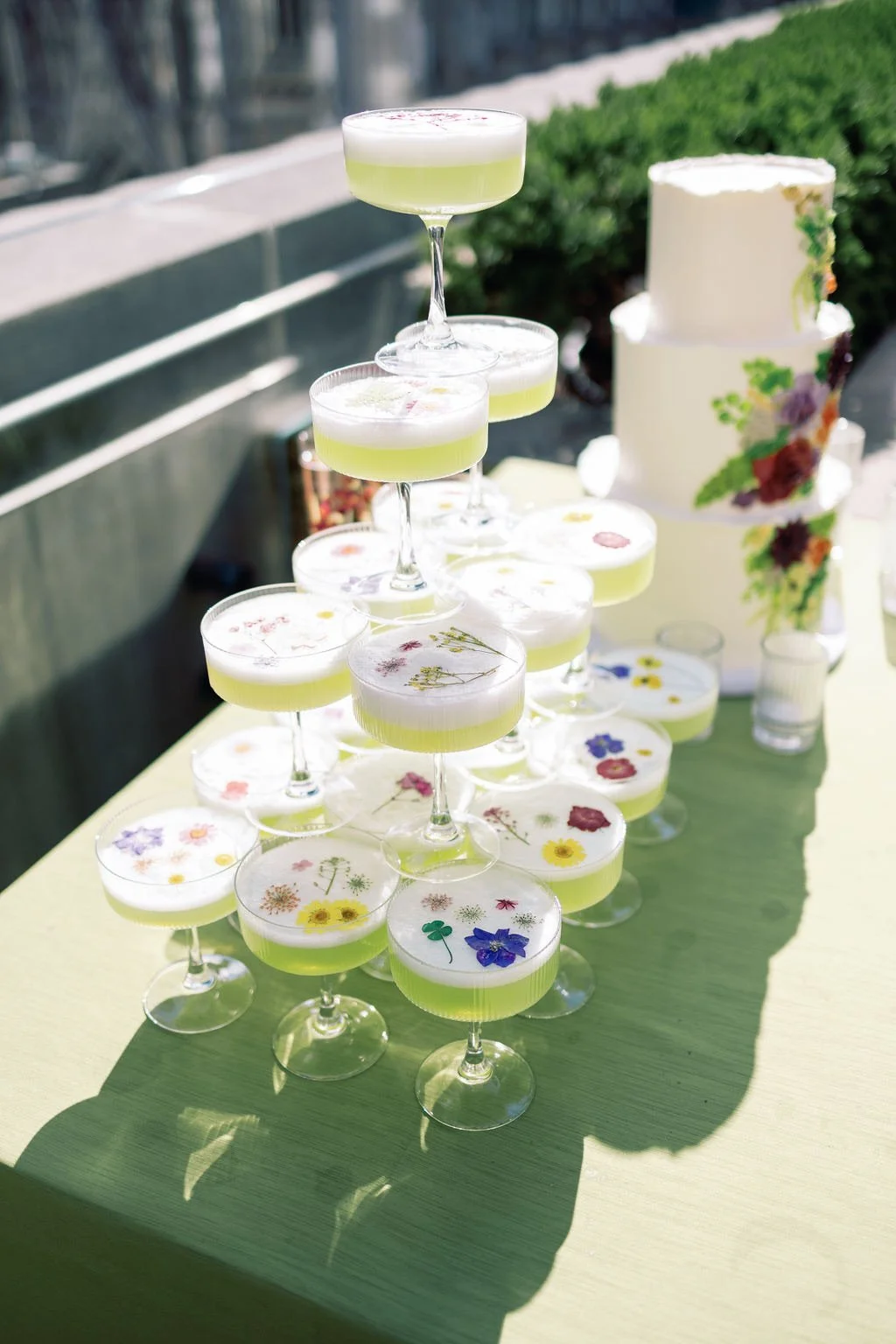 A pyramid of cocktail glasses filled with greenish-yellow drinks topped with foam, decorated with edible flowers, set on a table outdoors amidst a floral-themed cake and greenery.